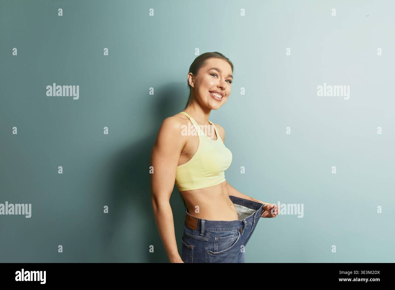 Eine Frau steht in einem hellen Raum mit einer blauen Wand und lächelt, während sie ihre lose Jeans hochhält. Sie hat eine Gewichtsreduktion erreicht und feiert ihren Erfolg mit Stockfoto