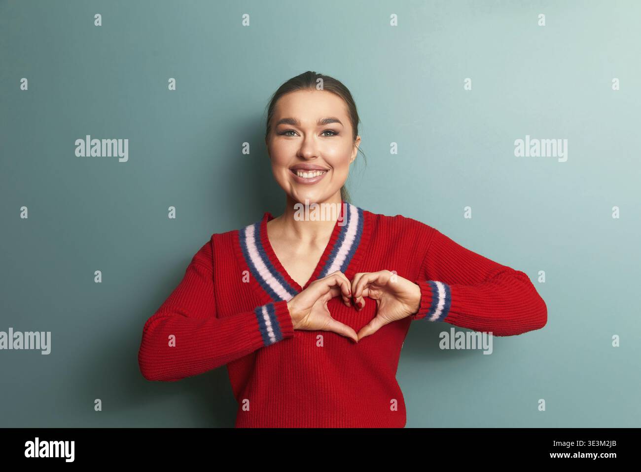Eine Frau steht vor einer grünen Wand und macht mit ihren Händen eine Herzform. Sie lächelt weit und trägt einen roten Pullover mit Streifen. Die Einstellung ist Stockfoto