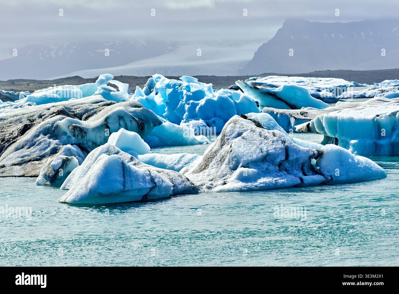 Eisberge schweben in den Gewässern der Jokulsarlon Glacier Lagoon. Die Aussicht zeigt eine Mischung aus Eis und ruhigem Wasser unter einem bewölkten Himmel. Stockfoto