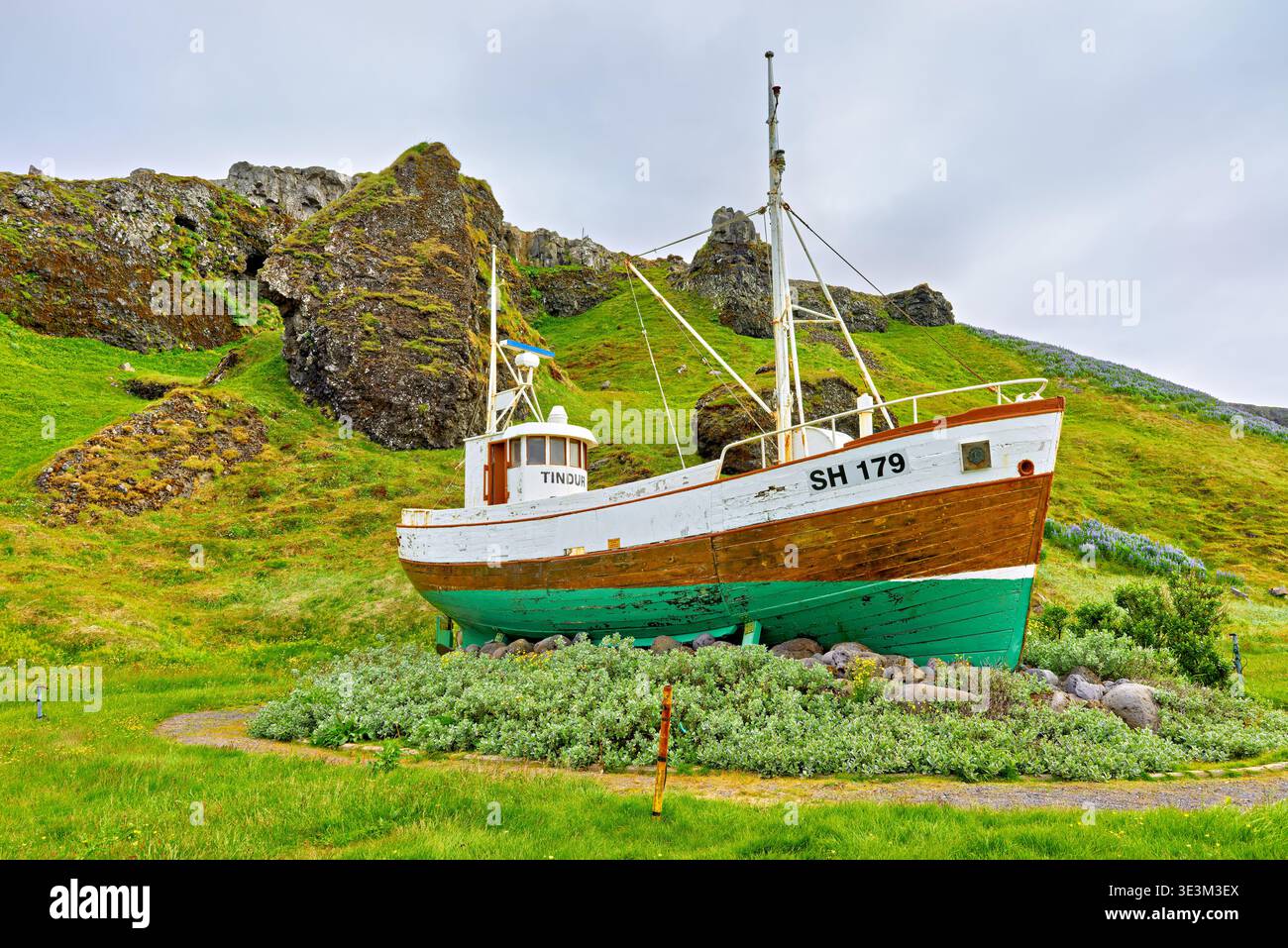 Ein Fischerboot ruht an Land in der Nähe von Olafsvik in Island. Gras und Felsen umgeben das Boot. Der Himmel ist bedeckt und grau. Stockfoto