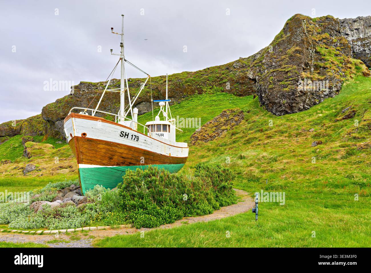 Ein Fischerboot steht an Land in Olafsvik, Island, umgeben von grünem Gras und Felsen. Das Gebäude scheint verwittert und verlassen zu sein. Stockfoto
