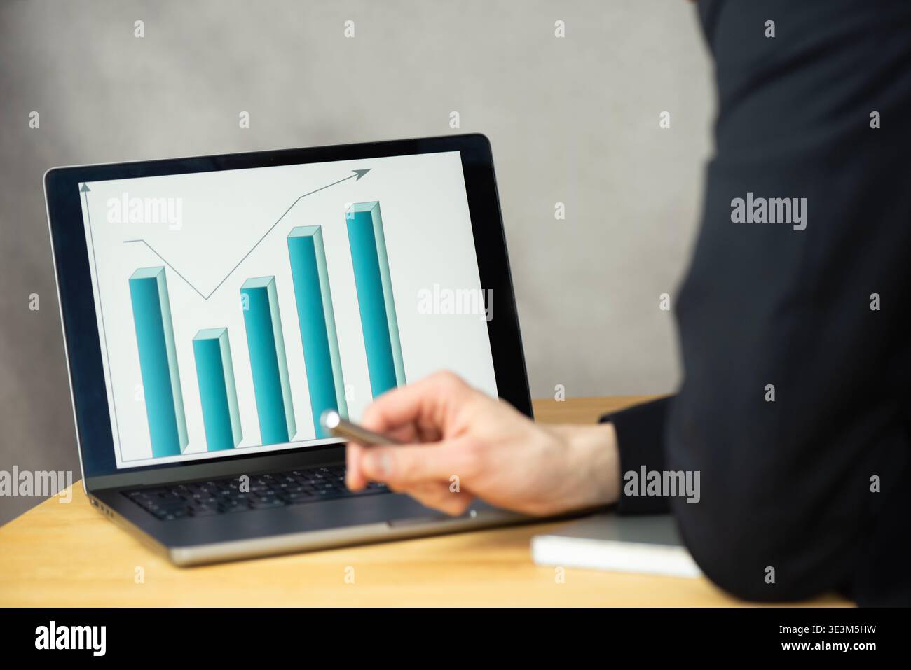 Ein Mann schaut auf einen Laptop, der ein Balkendiagramm anzeigt. Er hält einen Stift in der rechten Hand. Der Laptop steht auf einem Tisch. Stockfoto