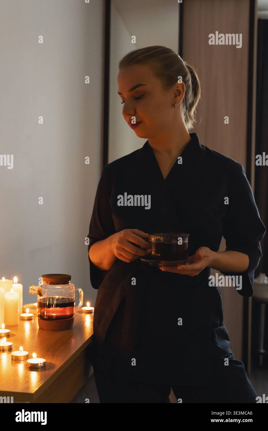 Frau gießt heißen Kräutertee in eine Glasschale mit aufsteigendem Dampf, umgeben von leuchtenden Kerzen, und schafft einen Moment des Wohlbefindens und Wohlbefindens Stockfoto