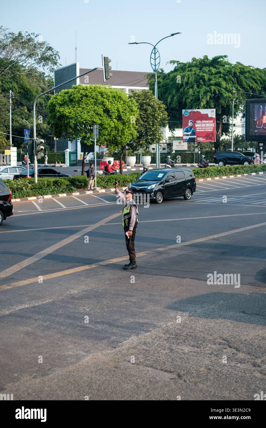 Fußgänger überquert die Straße an der Kreuzung. Der Verkehr fließt um den Quersteg herum. Grüne Bäume säumen den Straßenbereich. Moderne Straßenlaternen beleuchten die Szene. Stockfoto
