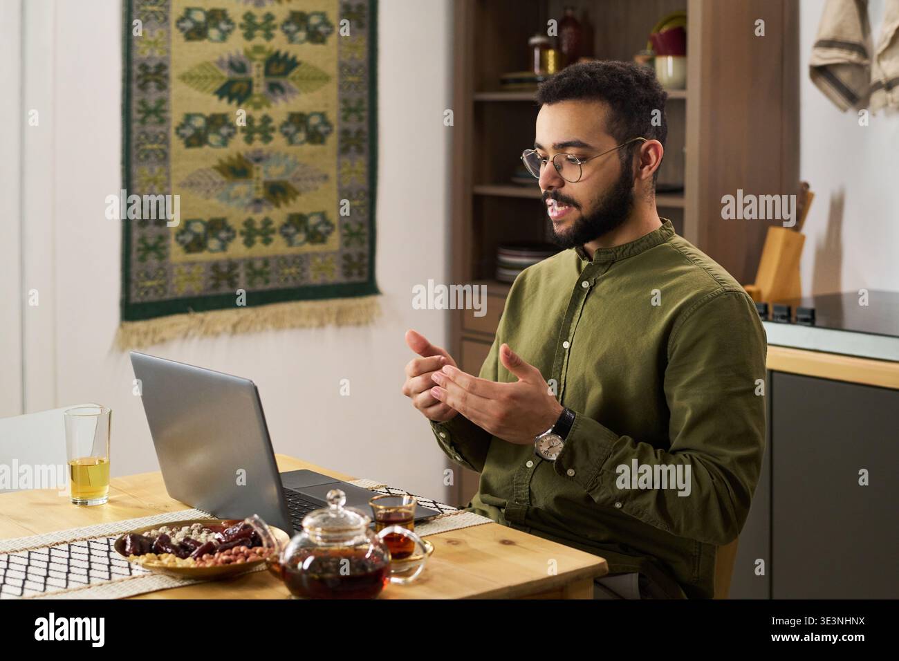 Junger erwachsener Mann aus dem Nahen Osten, der am Tisch sitzt, mit einem Laptop die Hände hebt, traditionelle Teesorte und Datteln auf dem Tisch, islamisches Dekor sichtbar im Hintergrund Stockfoto