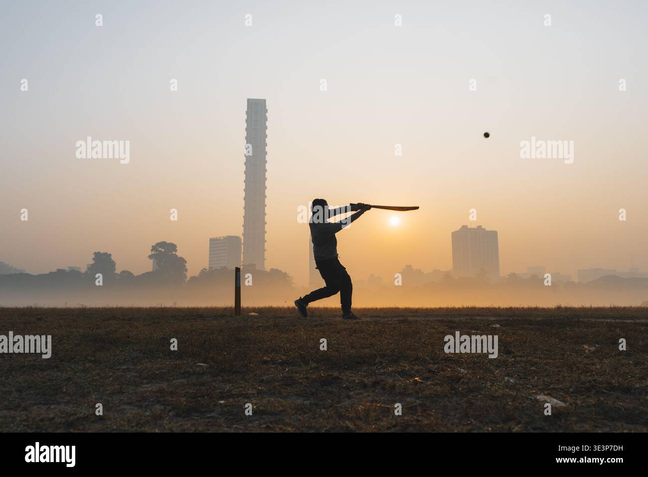 Blick auf einen einsamen Cricketspieler, der sich vor dem warmen Glanz der aufgehenden Sonne befindet und einen Schuss mit der Stadtlandschaft im nebeligen Hintergrund spielt, Kalkutta, West Ben Stockfoto