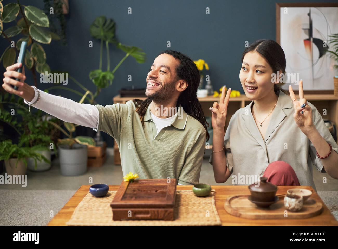Junger erwachsener Schwarzer Mann, der Selfie mit Smartphone macht, während er neben der jungen erwachsenen asiatischen Frau lächelt, die Friedenszeichen macht, beide sitzen am Tisch mit traditionellem Tee-Set Stockfoto
