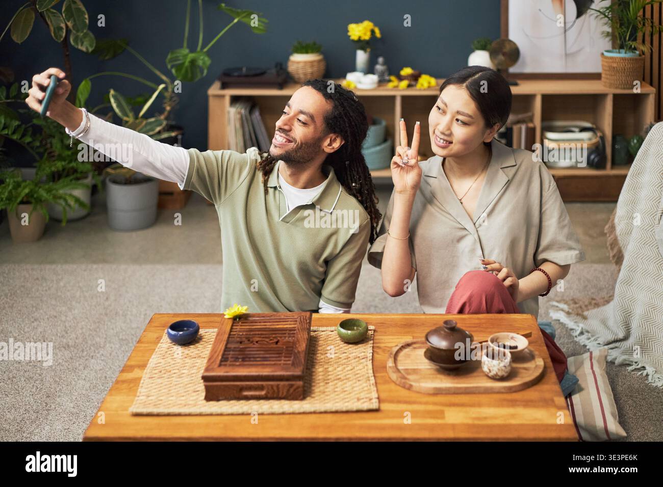 Junge Erwachsene asiatische Frau und junger Erwachsener Schwarzer Mann sitzen zusammen am niedrigen Tisch und machen Selfie mit Smartphone, asiatische Frau macht Friedenszeichen Geste, traditioneller Tee auf Tisch Stockfoto