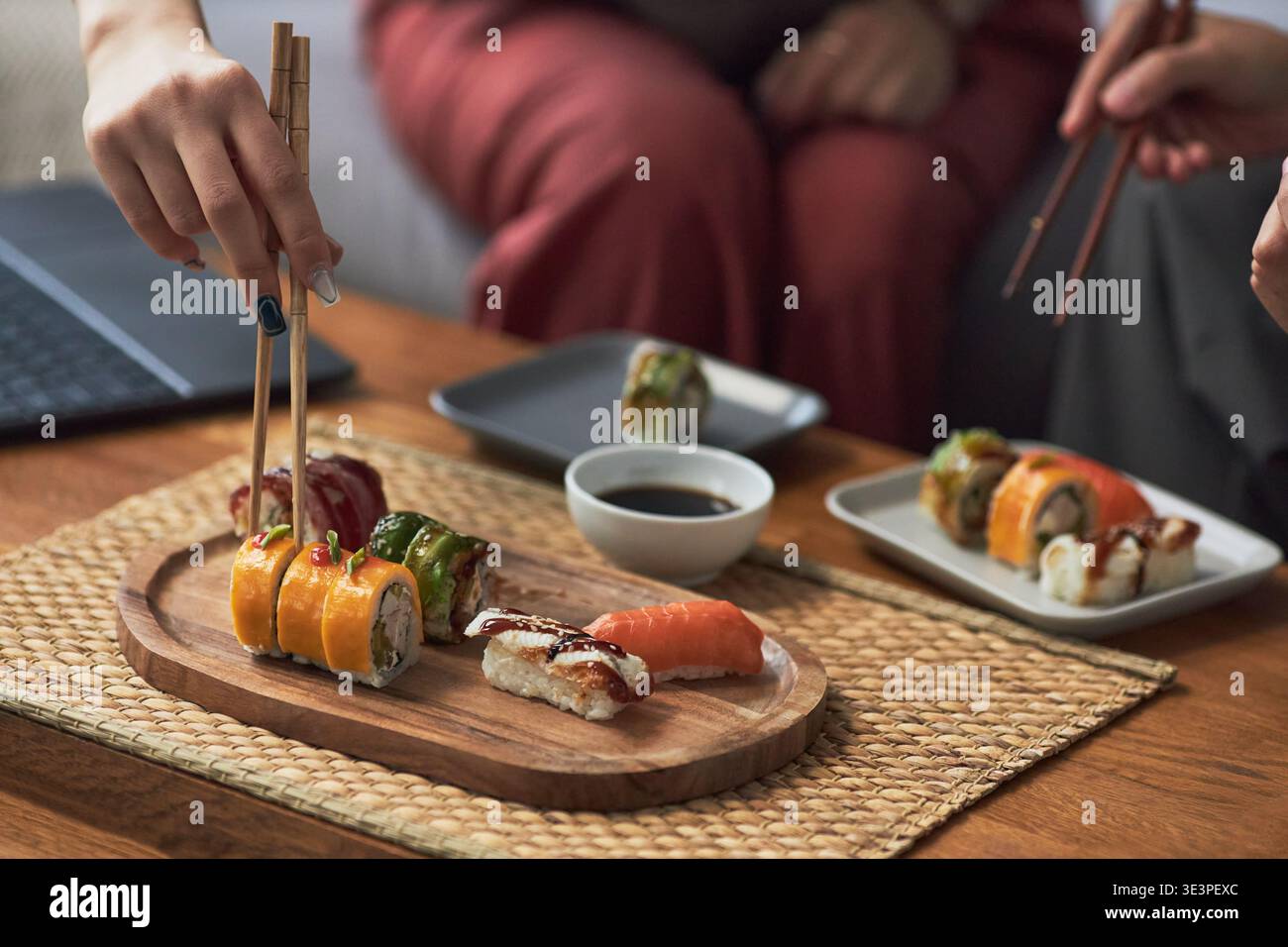 Asiatische junge Erwachsene pflücken verschiedene Sushi-Brötchen aus Holzplatte, teilen traditionelle japanische Gerichte während einer ungezwungenen Mahlzeit, Sojasauce in einem kleinen Gericht auf dem Tisch Stockfoto