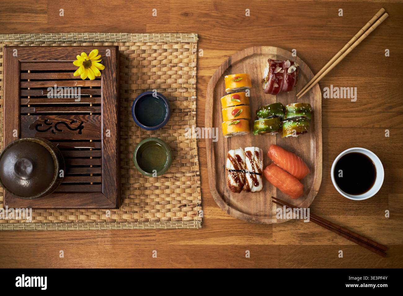 Traditionelle asiatische Küche mit verschiedenen Sushi-Stücken, auf Holzplatte mit Sojasauce und Essstäbchen neben Tee auf gewebter Matte, die kulturelle Elemente hervorheben Stockfoto