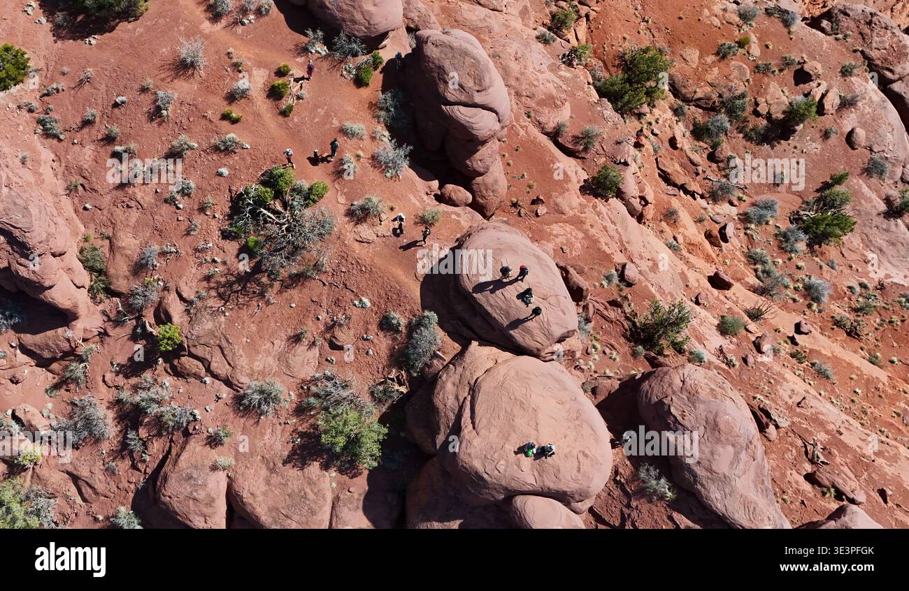 Aus der Vogelperspektive rostfarbene Felsen und karge Vegetation zeichnen eine zerklüftete Landschaft, in der winzige Figuren die Weite von Moab, Utah, USA, erkunden. Stockfoto