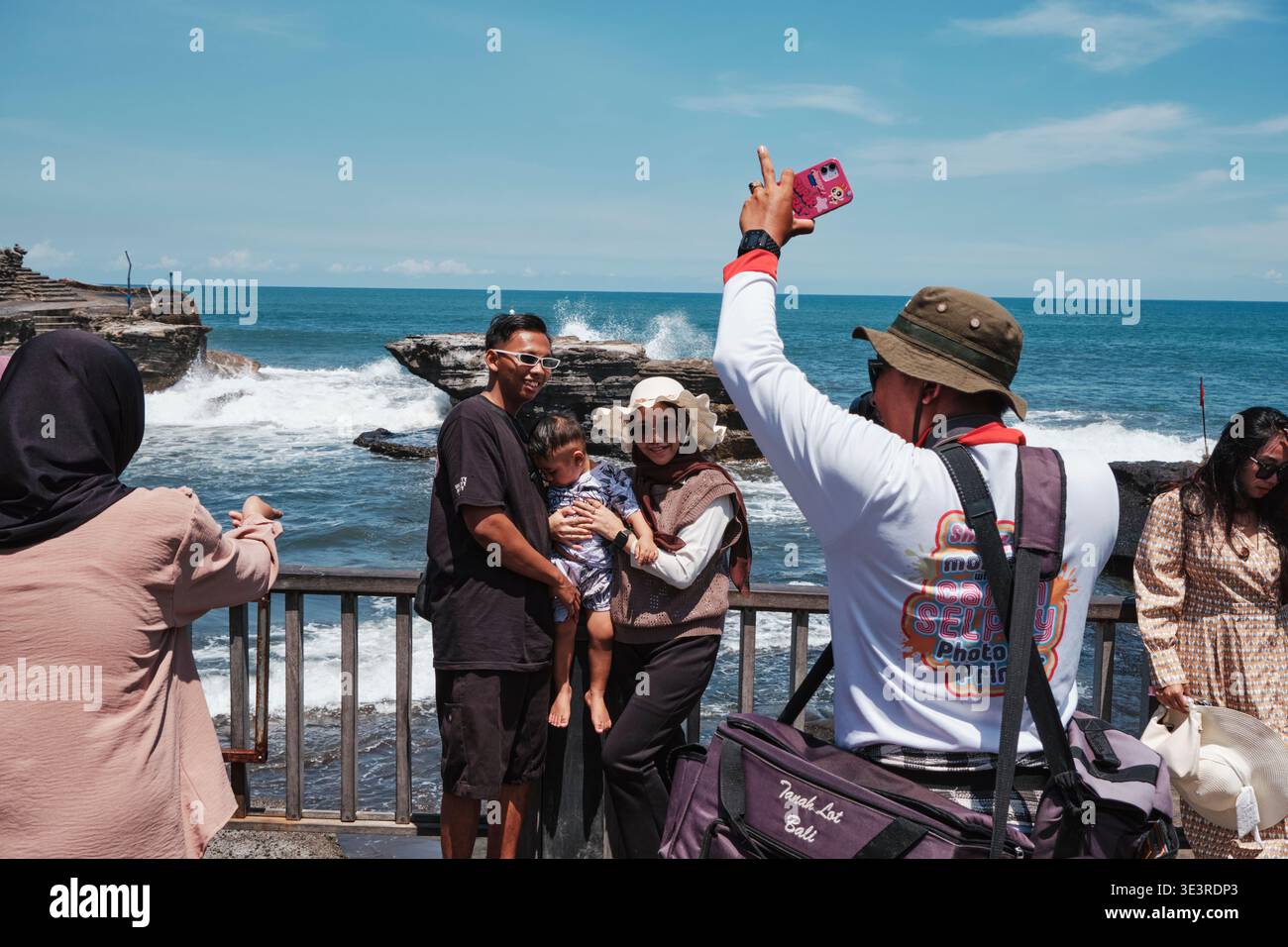 Gruppen von Menschen genießen einen Tag im Tanah Lot auf Bali. Familien fotografieren zusammen mit dem Meer als Kulisse. Die Sonne scheint hell. Stockfoto