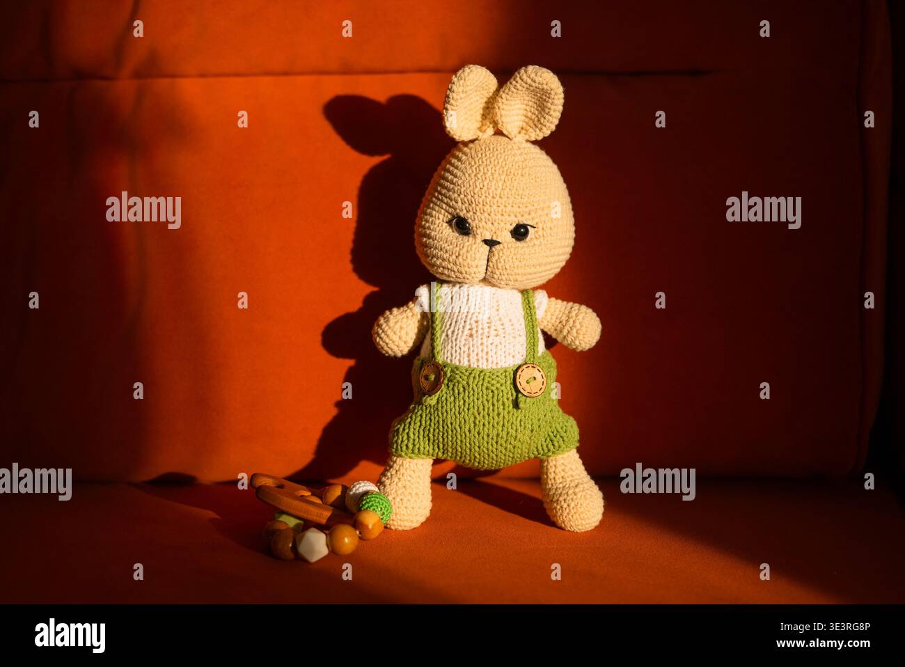 Amigurumi Hase Plüschpuppe in grün gestrickter Latzhose, die auf einer warmen orangefarbenen Couch steht und Themen wie handgefertigtes Spielzeug, Kinderspiel, Basteln und Kinderzimmer-Dekoration illustriert Stockfoto