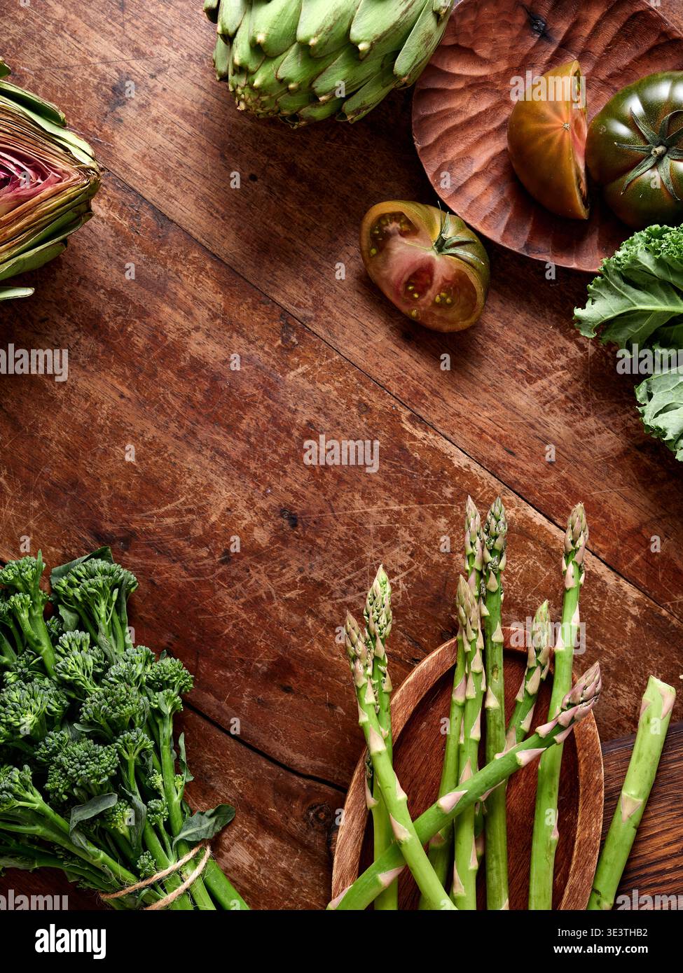 Frisches Gemüse wird auf einem Holztisch serviert. Die Szene zeigt Brokkoli, Spargel, Artischocken und Tomaten. Diese Einstellung schlägt eine Mahlzeit vor Stockfoto