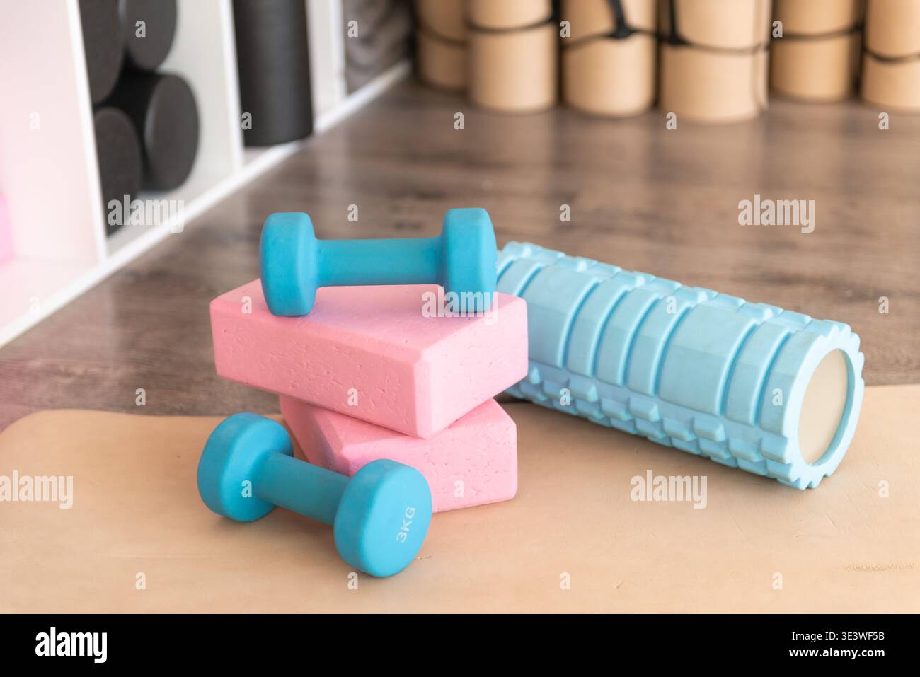 Ein Satz blauer Kurzhanteln, rosafarbener Yoga-Blöcke und eine strukturierte Schaumstoffrolle sind ordentlich auf einer Yoga-Matte in einem Fitnessstudio angeordnet und fördern so Fitness und Wohlbefinden im Fitnessstudio Stockfoto