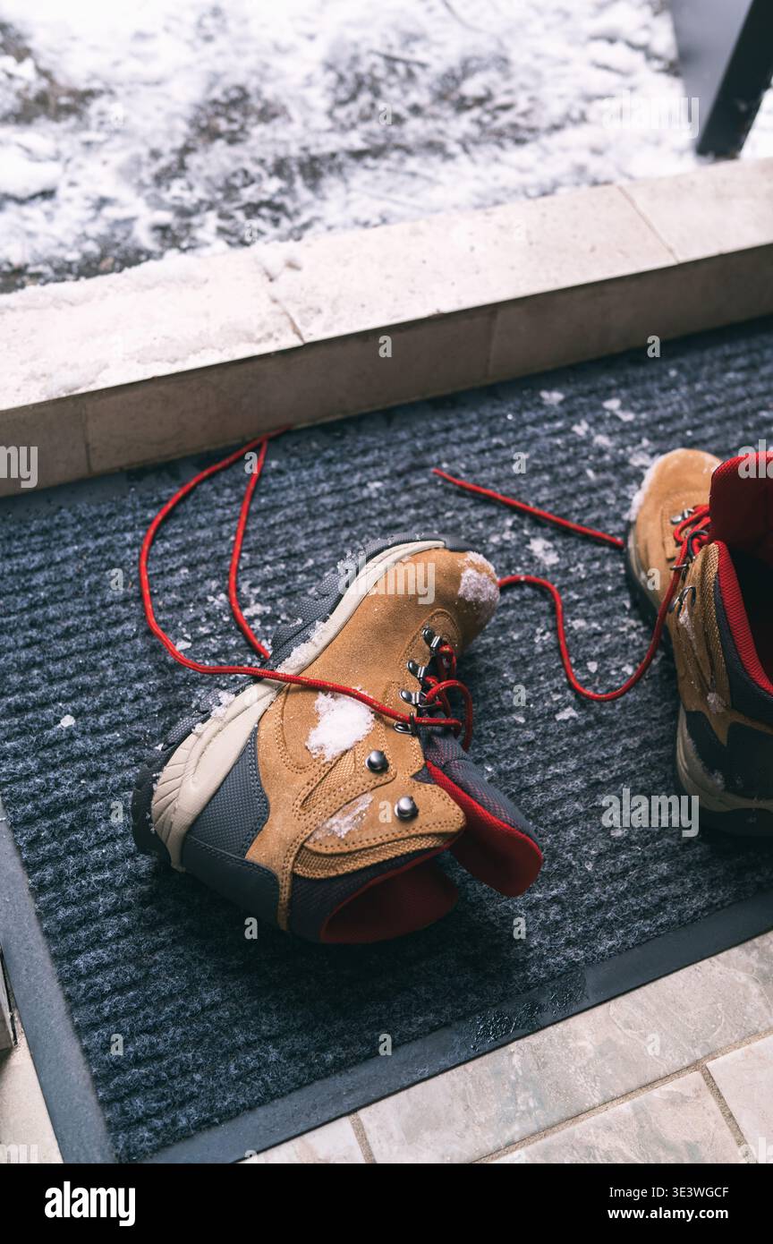 Ein Paar robuste Winterstiefel mit roten Schnürsenkeln liegt auf einer schneebedeckten Fußmatte. Die Szene fängt das Wesen des kalten Wetters ein und betont Wärme A Stockfoto