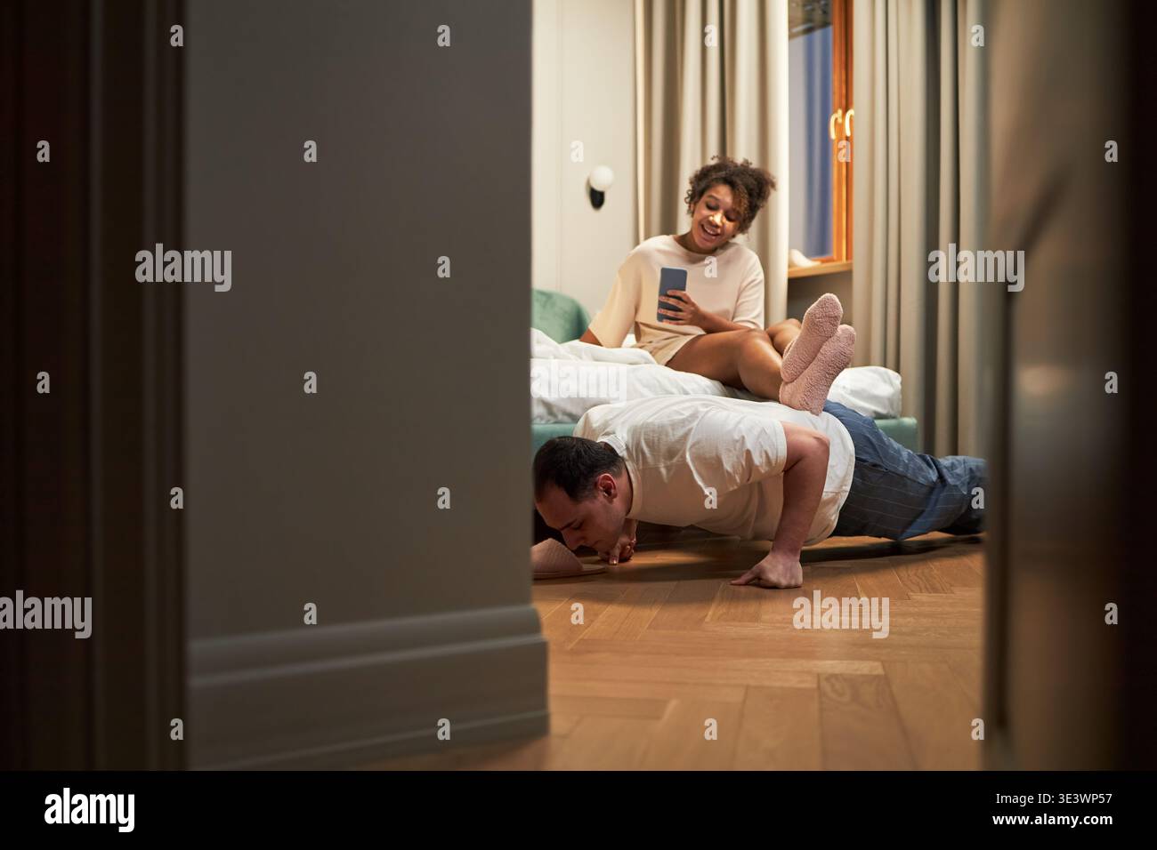 Kaukasischer Mann mittleren Alters, der Liegestütze auf Holzboden macht, während Schwarze junge Erwachsene Frau auf dem Bett sitzt, Smartphone hält und lacht, beide in spielerischer Interaktion im modernen Schlafzimmer Stockfoto