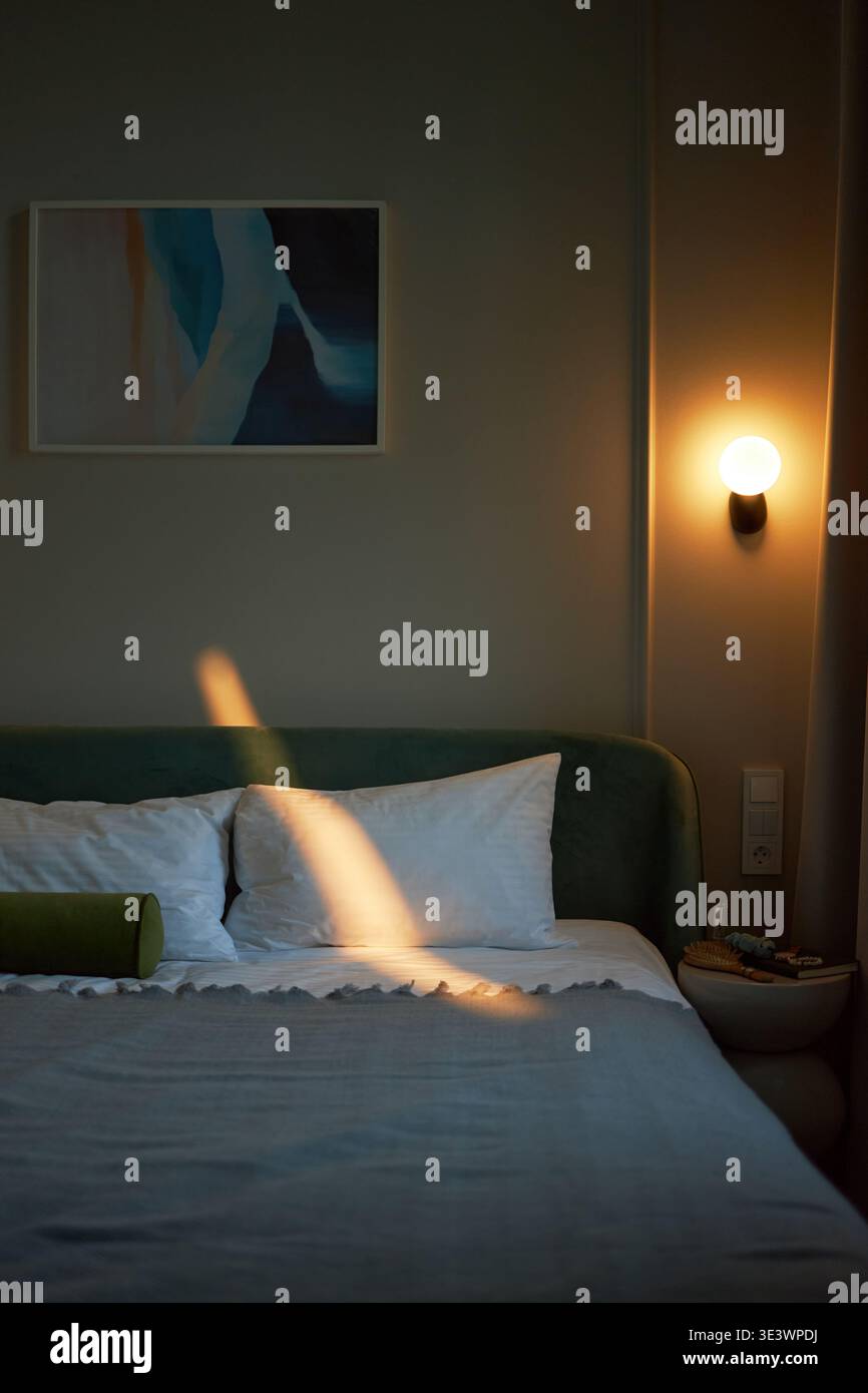 Vertikal des leeren, modernen Schlafzimmers mit ordentlich gefertigtem Bett mit Kissen, abstrakter Malerei an der Wand, beleuchteter Wandleuchte, die warmes Licht ausstrahlt, keine Menschen sichtbar, ruhige Inneneinrichtung Stockfoto
