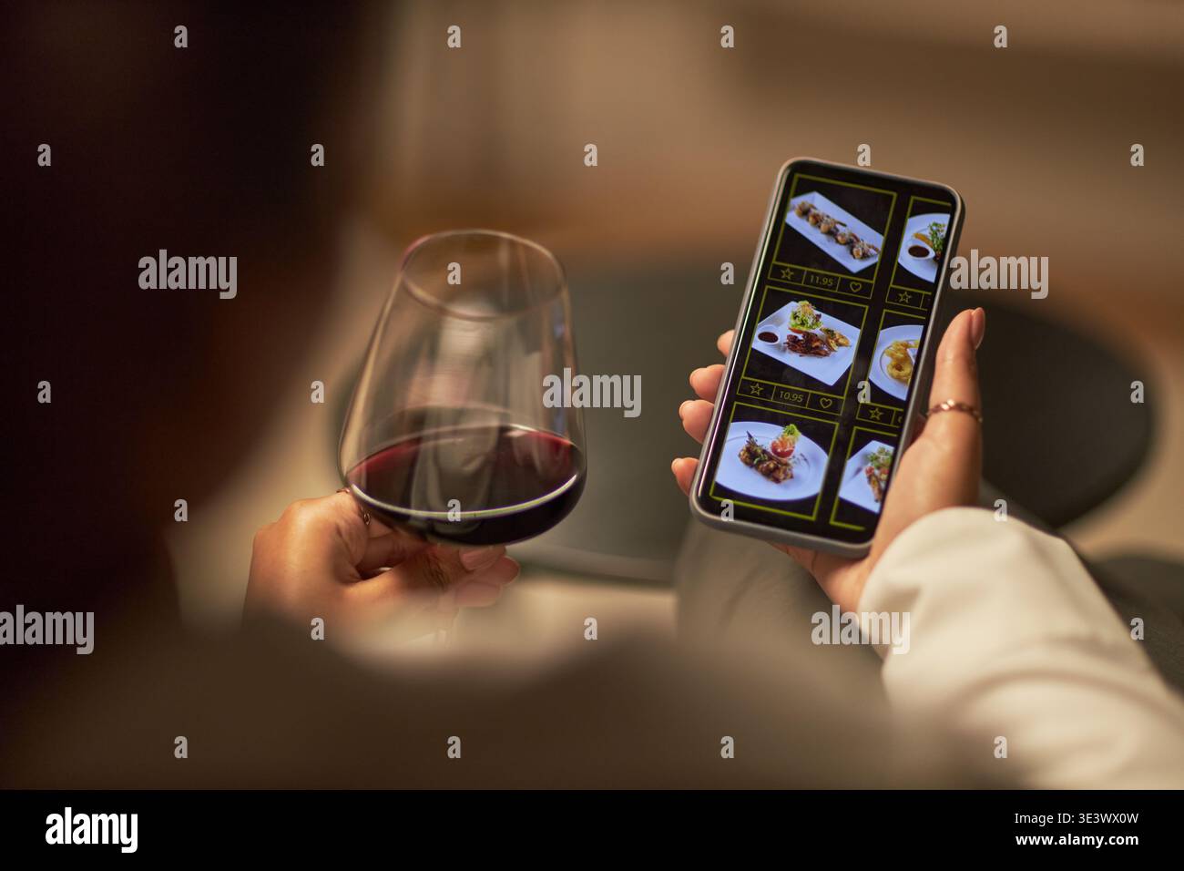 Kaukasische junge Erwachsene Frau, die ein Glas Rotwein hält, während sie auf dem Smartphone das Restaurant-Menü mit Gourmetgerichten durchstöbert und drinnen sitzt, mit Fokus auf die Hände und den Bildschirm des Telefons Stockfoto