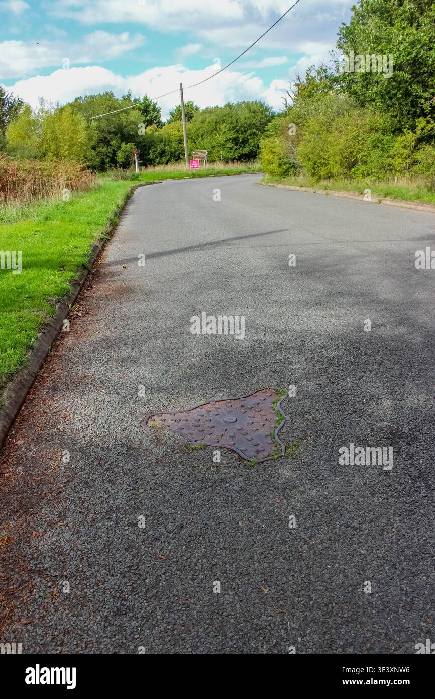 Eine Asphaltstraße im Vereinigten Königreich führt durch eine üppige grüne Landschaft mit einem markanten dreieckigen Mannloch im Vordergrund. Stockfoto