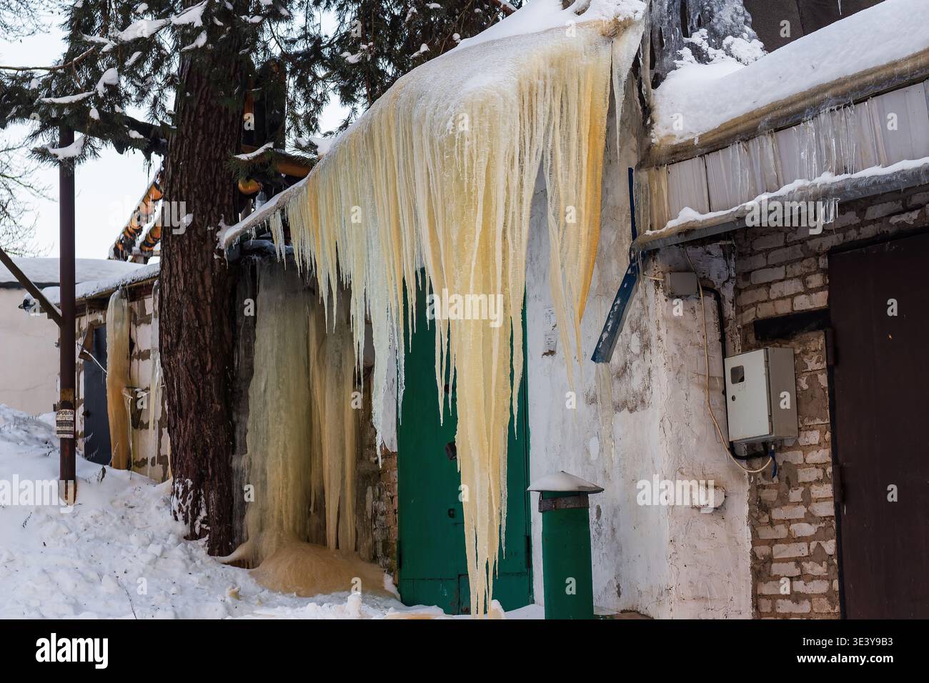 Riesige, gelbliche Eiszapfen hängen dramatisch von alten Garagendächern und verwandeln eine banale Winterszene in ein bezauberndes, gefrorenes Naturspektakel. Stockfoto