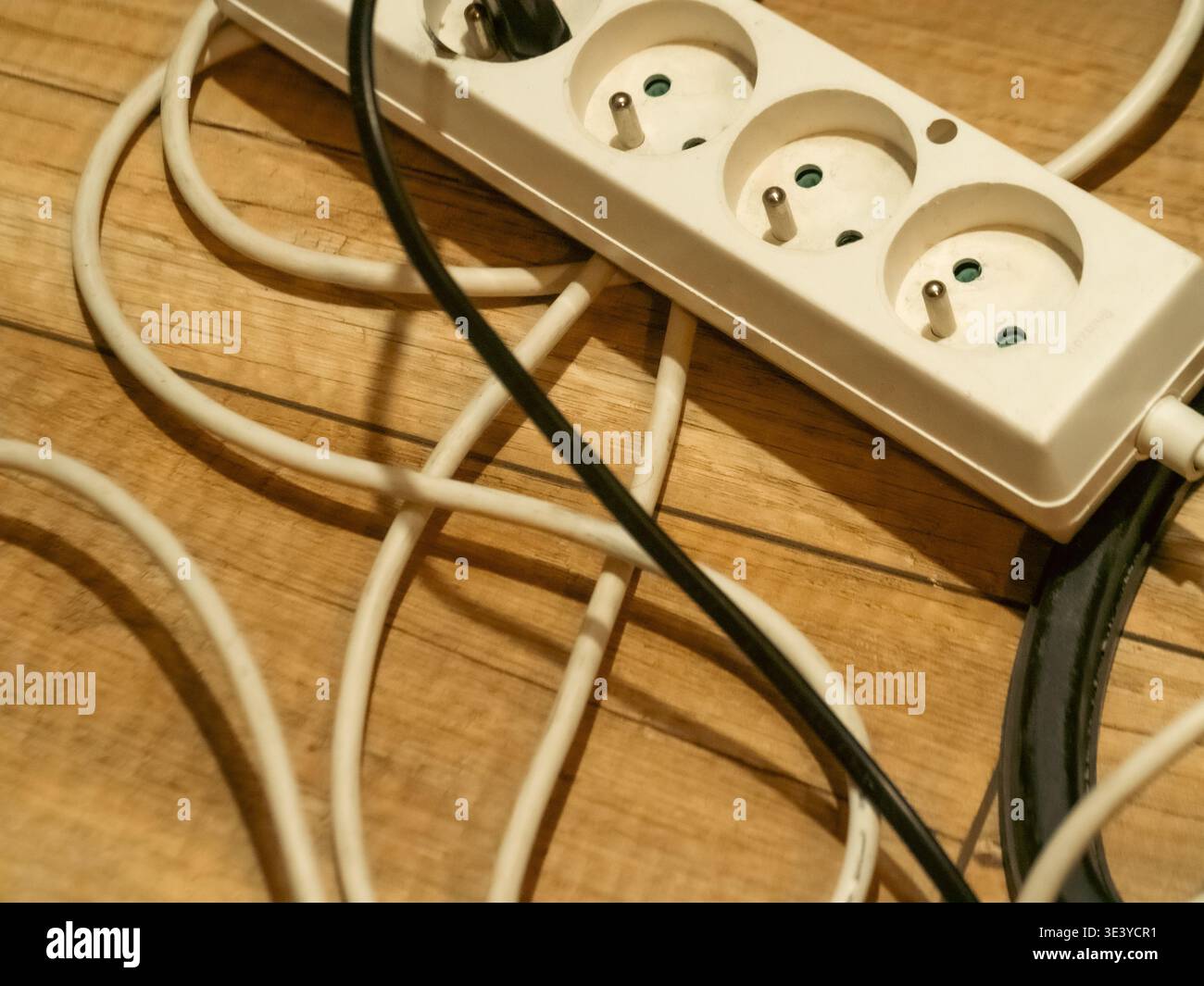 Verwickelte Kabel umgeben eine beige Steckdosenleiste auf Holzboden. Die Kabel spulen sich locker und zeigen eine unordentliche elektrische Einrichtung. Steckdosenleiste mit drei europäischen Stk Stockfoto