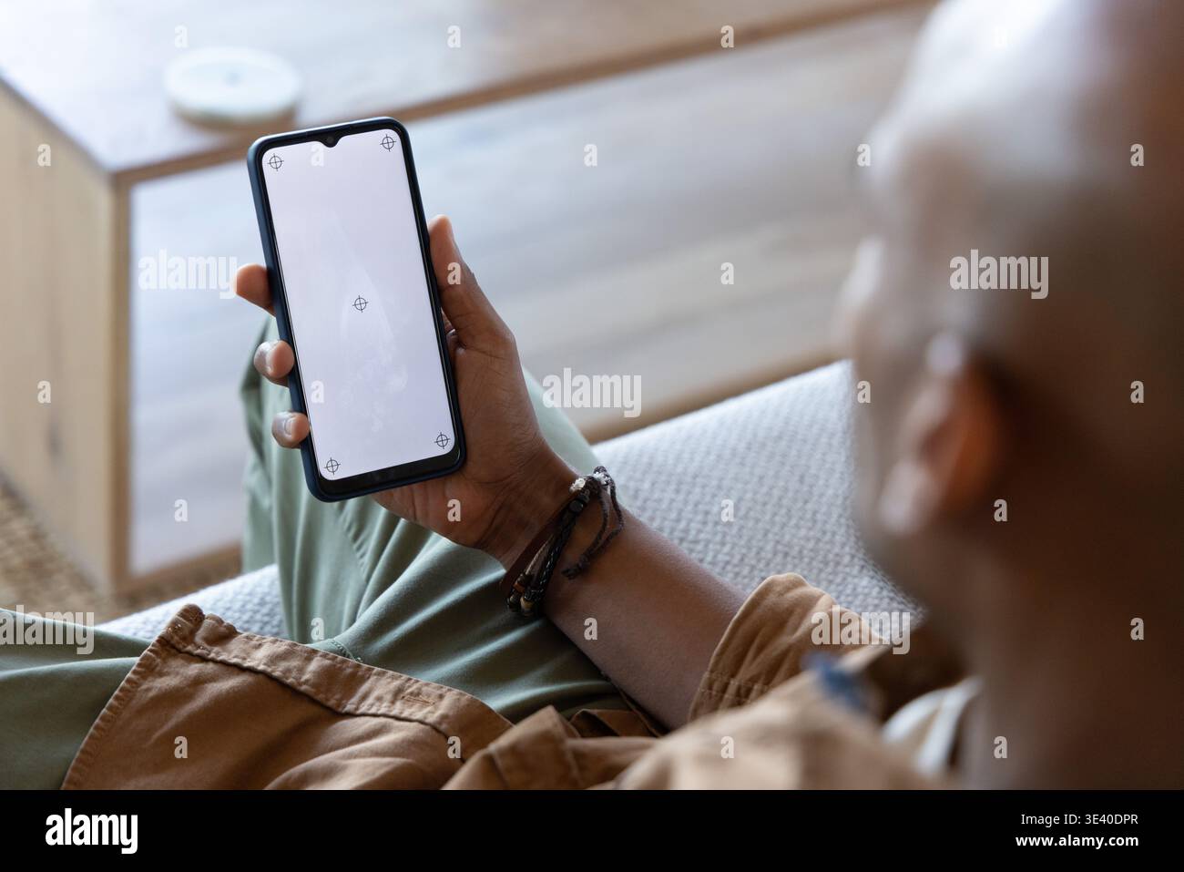 Afroamerikaner sitzt auf dem Sofa und hält Telefonmockup in braunem Hemd und Armbändern, Kopierraum Stockfoto