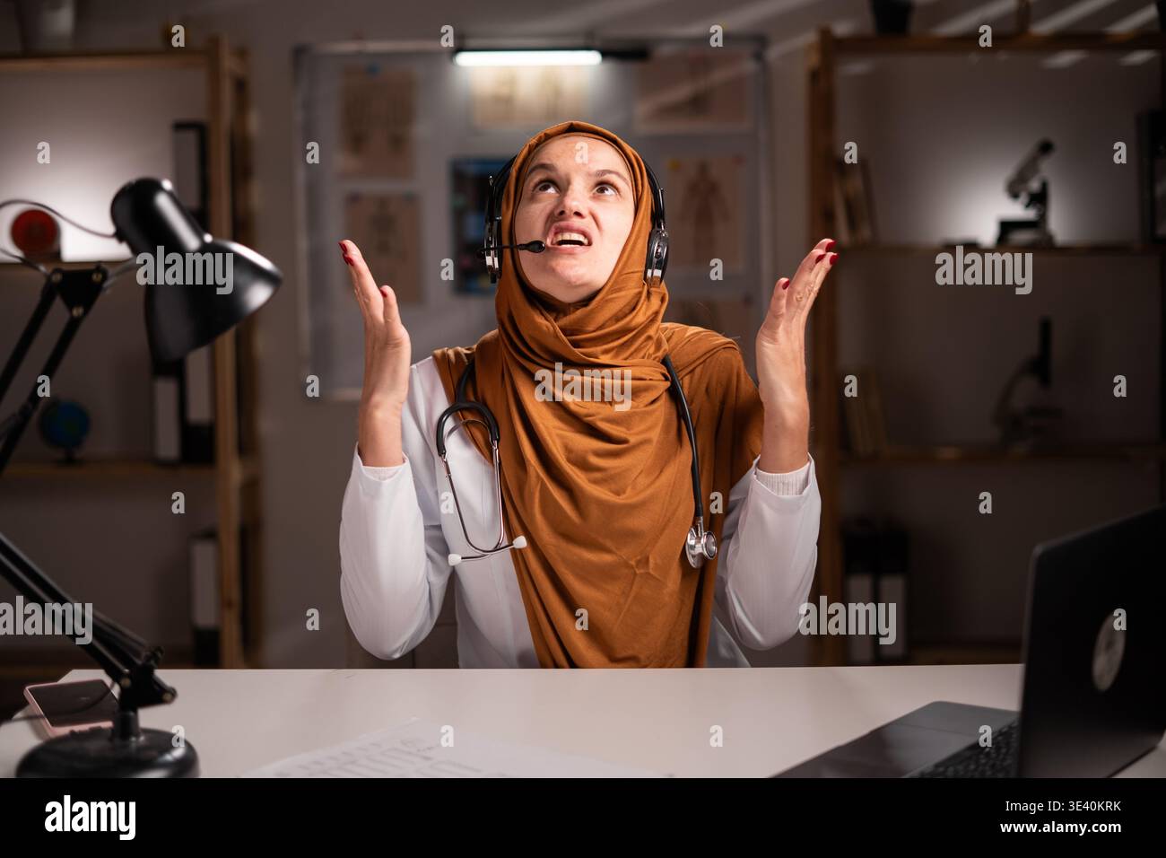 Verärgerte weibliche muslimische Ärztin im Hijab oder Arzttherapeutin im weißen Mantel Tragen Sie Headset mit Mikrofon arbeiten mit Laptop schreiende Hände nach oben während n Stockfoto