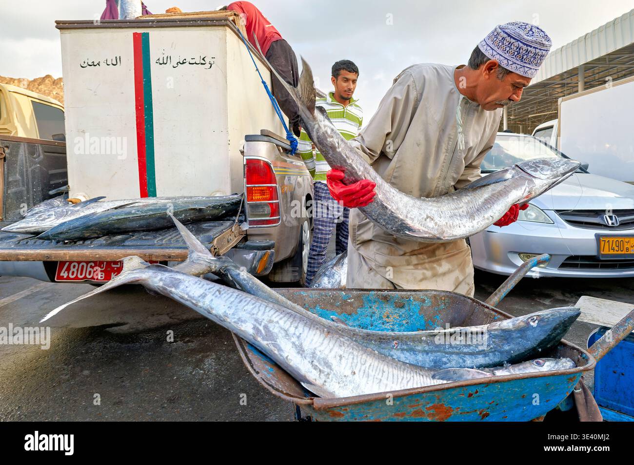Arbeiter transportieren große Fische auf dem Muscat-Fischmarkt. Die Szene zeigt beschäftigte Menschen und Fahrzeuge. Es ist tagsüber in Oman. Stockfoto