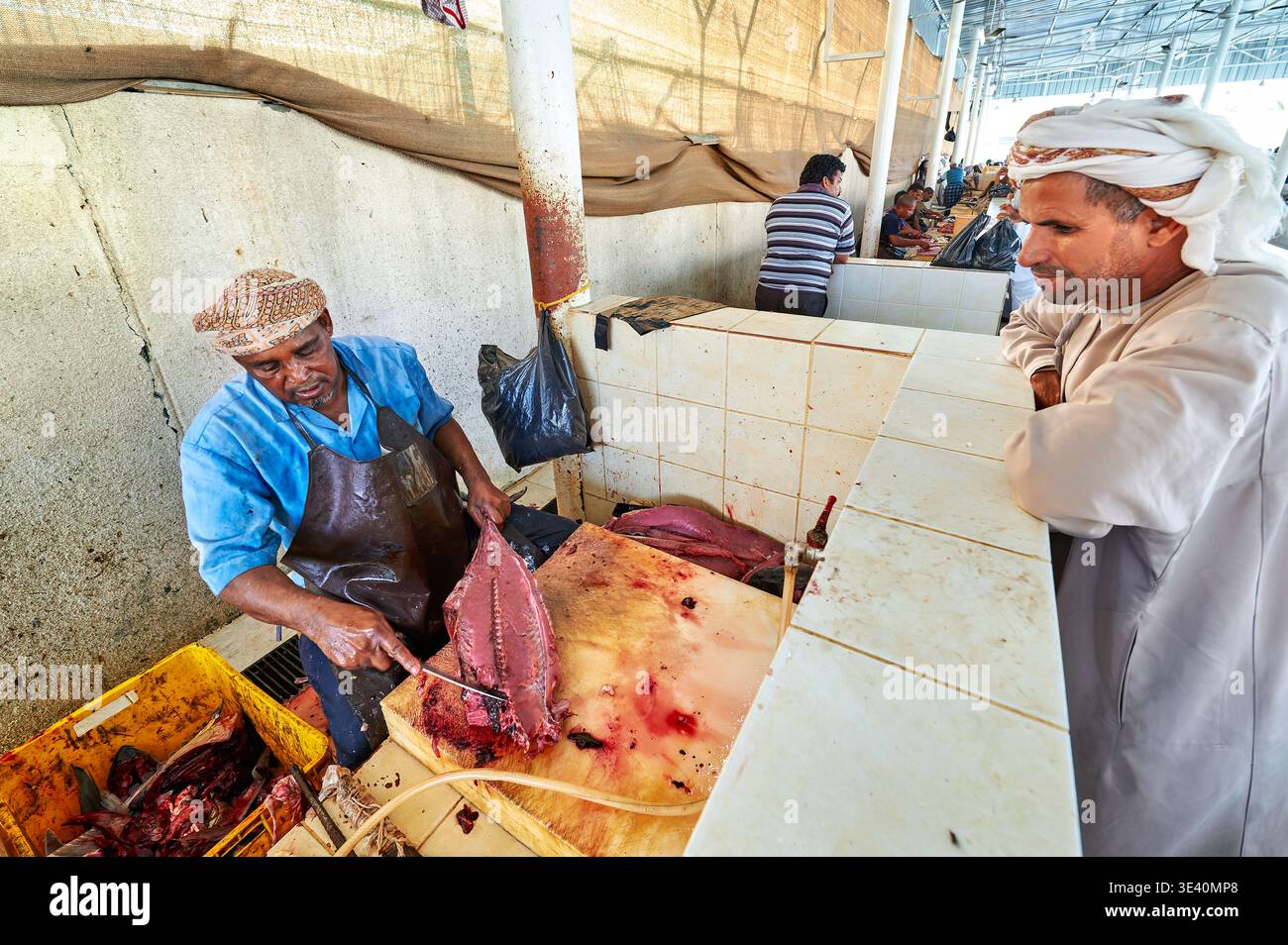 Fischhändler schneiden Fisch und bereiten ihn auf dem Markt in Maskat, Oman, zu. Kunden beobachten die Arbeit, die um sie herum stattfindet, während der Markt geschäftig ist. Stockfoto