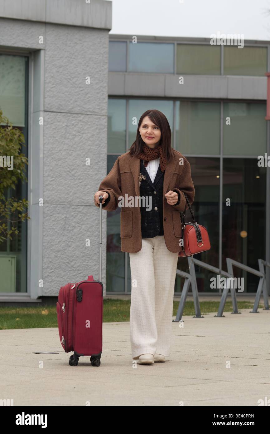Eine Frau bereitet sich auf eine Reise vor, steht mit Gepäck vor einem städtischen Gebäude. Das Foto spiegelt Komfort, Reife und zeitlose Mode wider. Stockfoto