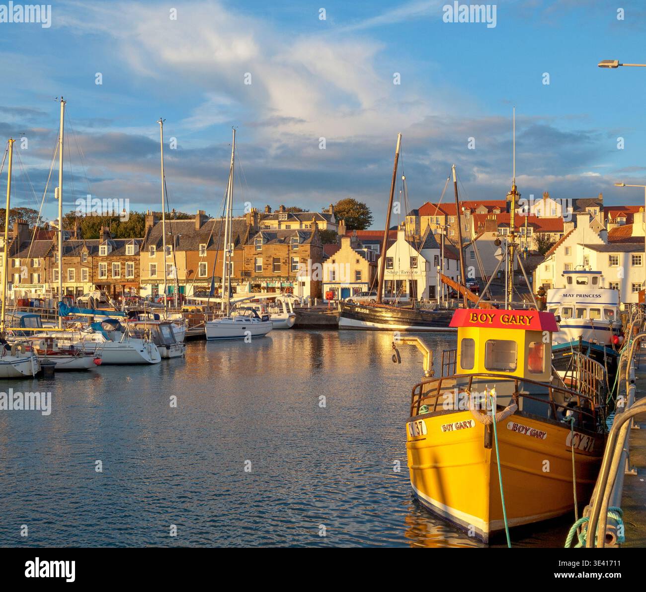 Der Hafen von Anstruther leuchtet in warmem goldenem Licht, während farbenfrohe Fischerboote neben einem Steinufer mit Geschäften am Ufer, Anlegejachten und traditionellen Stockfoto