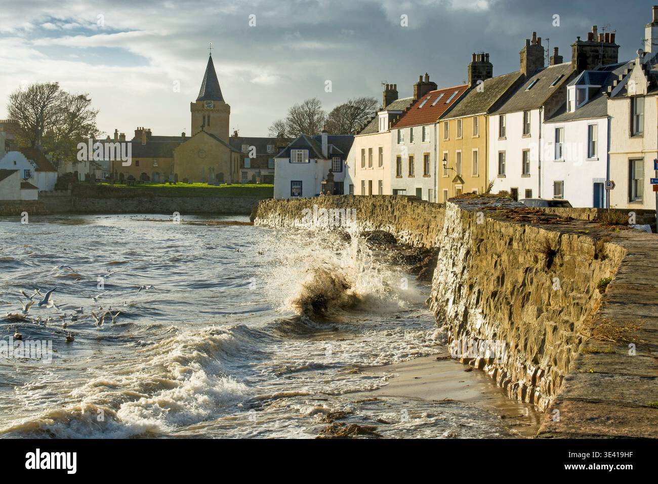Stürmische Wellen krachen gegen die Steinmauer in Anstruther, Fife, Schottland, und erleuchten das Küstendorf, den Kirchturm und die Reihen historischer Hohs Stockfoto