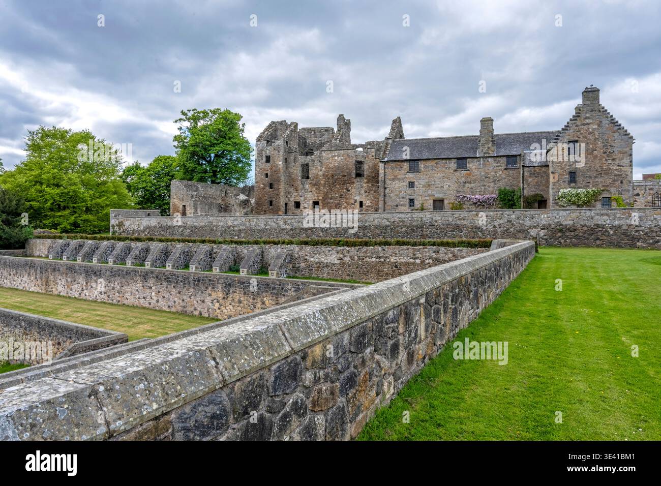 Historisches Aberdour Castle, eingerahmt von terrassierten Steinmauern, gepflegten Rasenflächen und Reifen Bäumen unter einem dramatischen bewölkten Himmel, das mittelalterliche Archite hervorhebt Stockfoto