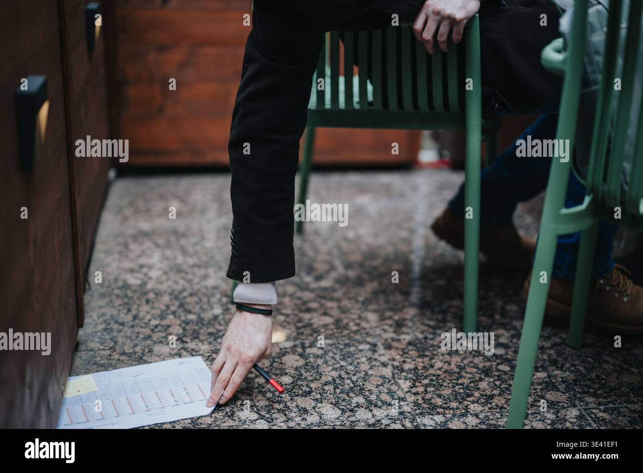 Ein Geschäftsmann greift, um ein Dokument und einen Stift unter einem Stuhl in einem Café im Freien abzuholen Stockfoto