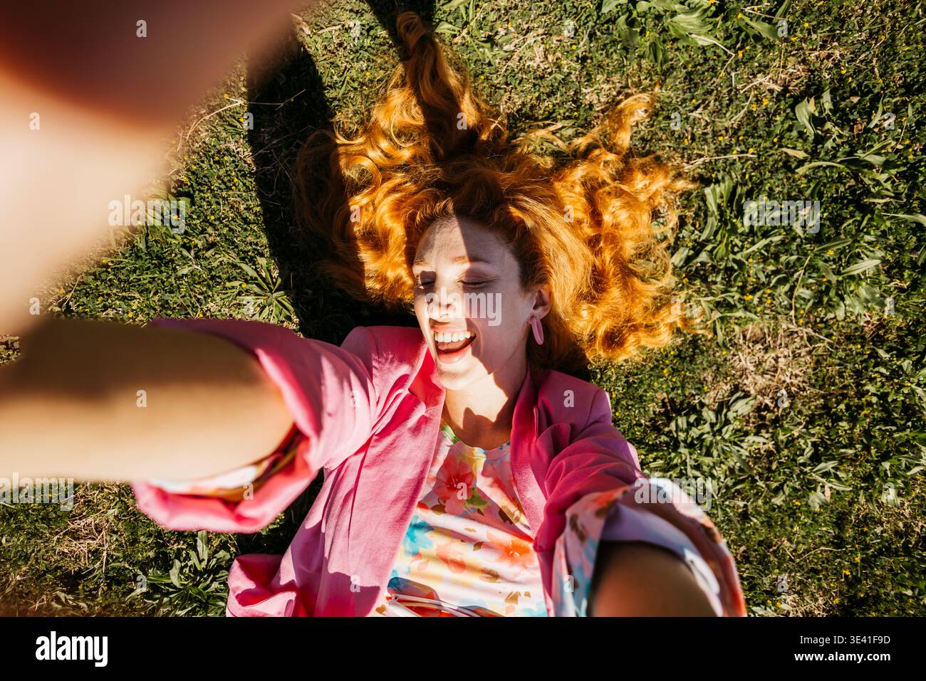 Lächelnde Frau mit rotem Haar, die auf grünem Gras liegt und an einem sonnigen Tag ein Selfie macht Stockfoto