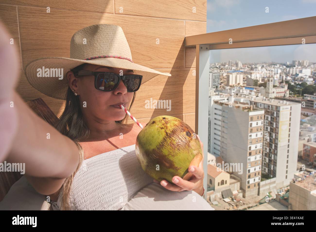 Frau, die sich auf einem Balkon entspannt und Kokoswasser mit Blick auf die Stadt trinkt Stockfoto