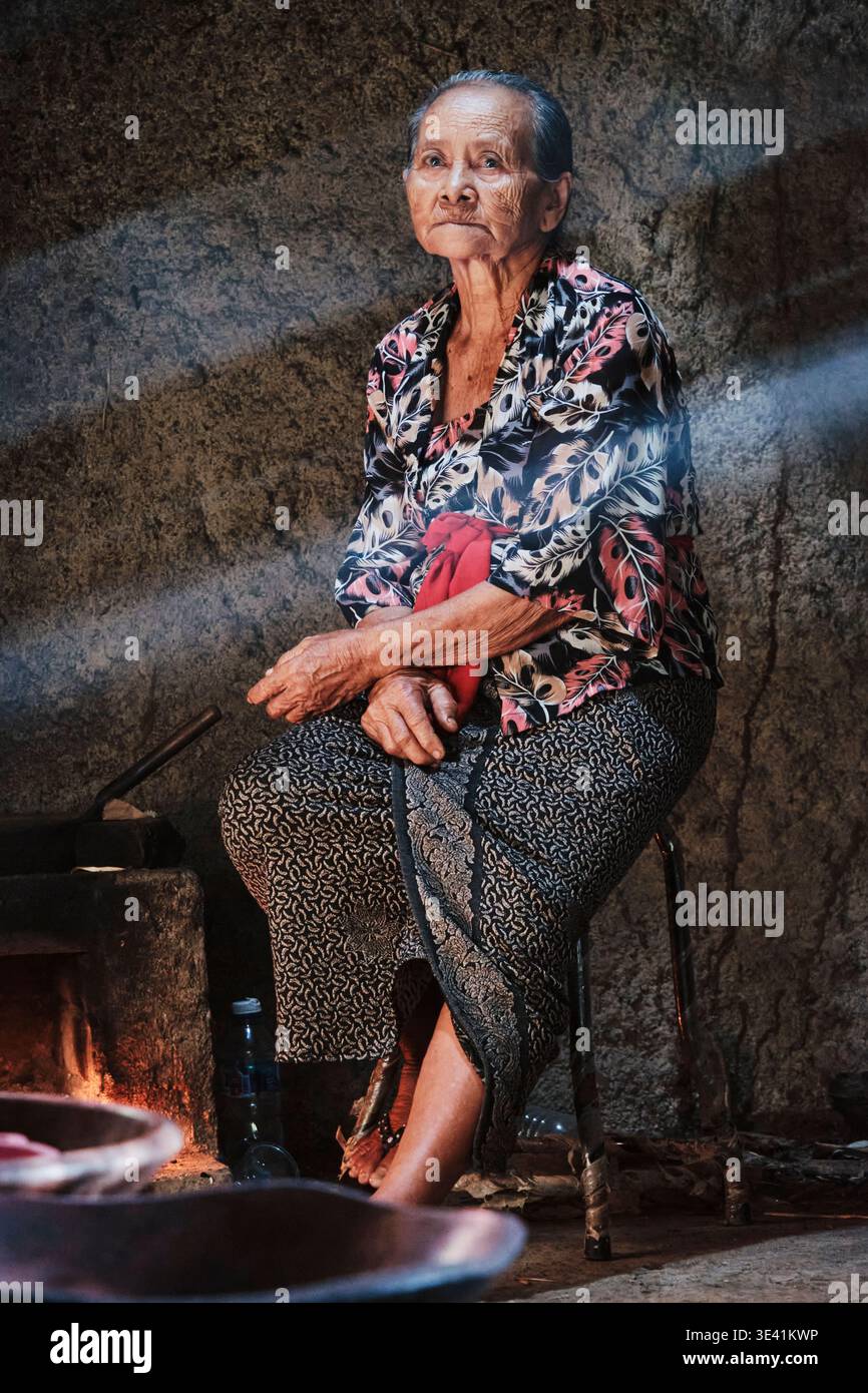 Eine ältere balinesische Frau sitzt auf einem Stuhl in einer einfachen Küche auf Bali. Das Morgenlicht leuchtet durch den Raum, während sie traditionelle Speisen zubereitet. Stockfoto