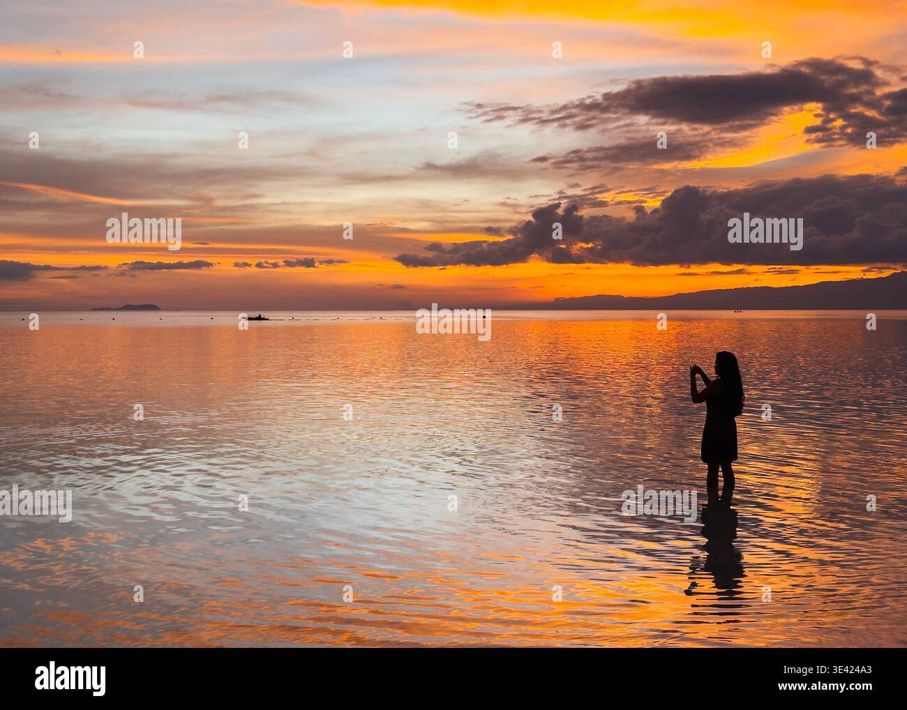 Silhouette einer Frau bei Sonnenuntergang am Little Paliton Beach, Siquijor, Philippinen, mit ruhigem Meer, das leuchtend orange und goldene Himmel reflektiert. Stockfoto