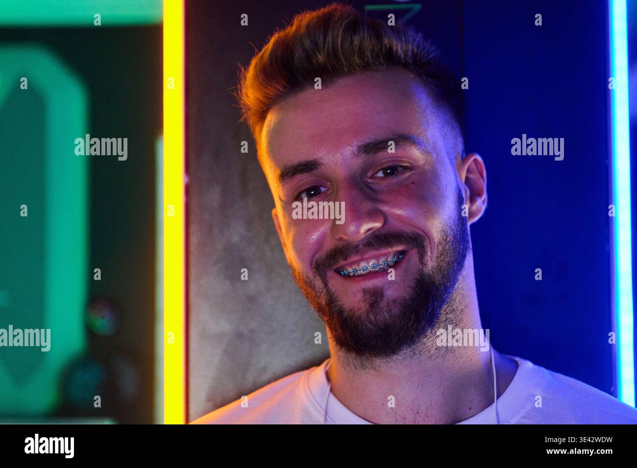 Junger Mann mit Zahnspangen lächelnd in Neon Porträt, lässigem Bart und farbenfroher LED-Beleuchtung Stockfoto