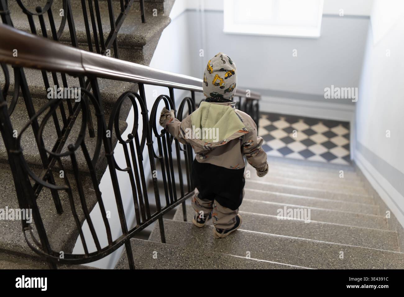 Rückansicht eines Kleinkindes, das eine Steintreppe im Appartementgebäude hinuntergeht, in saisonaler Kleidung, Heimroutine, Mobilitätsunterricht und Familiensafe gebündelt Stockfoto