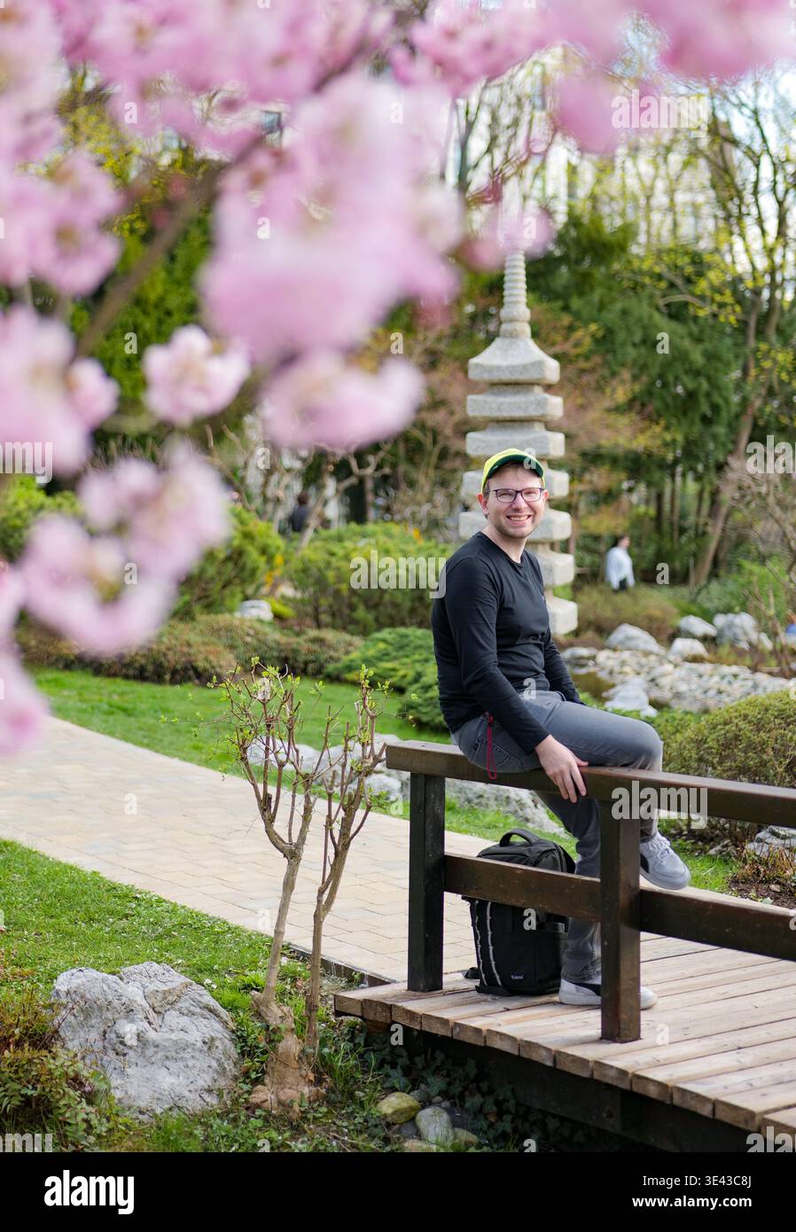 Ein lächelnder Mann sitzt auf einer Holzbrücke in einem japanischen Garten mit Kirschblüten. Frühes Erwachen der Natur. Stockfoto