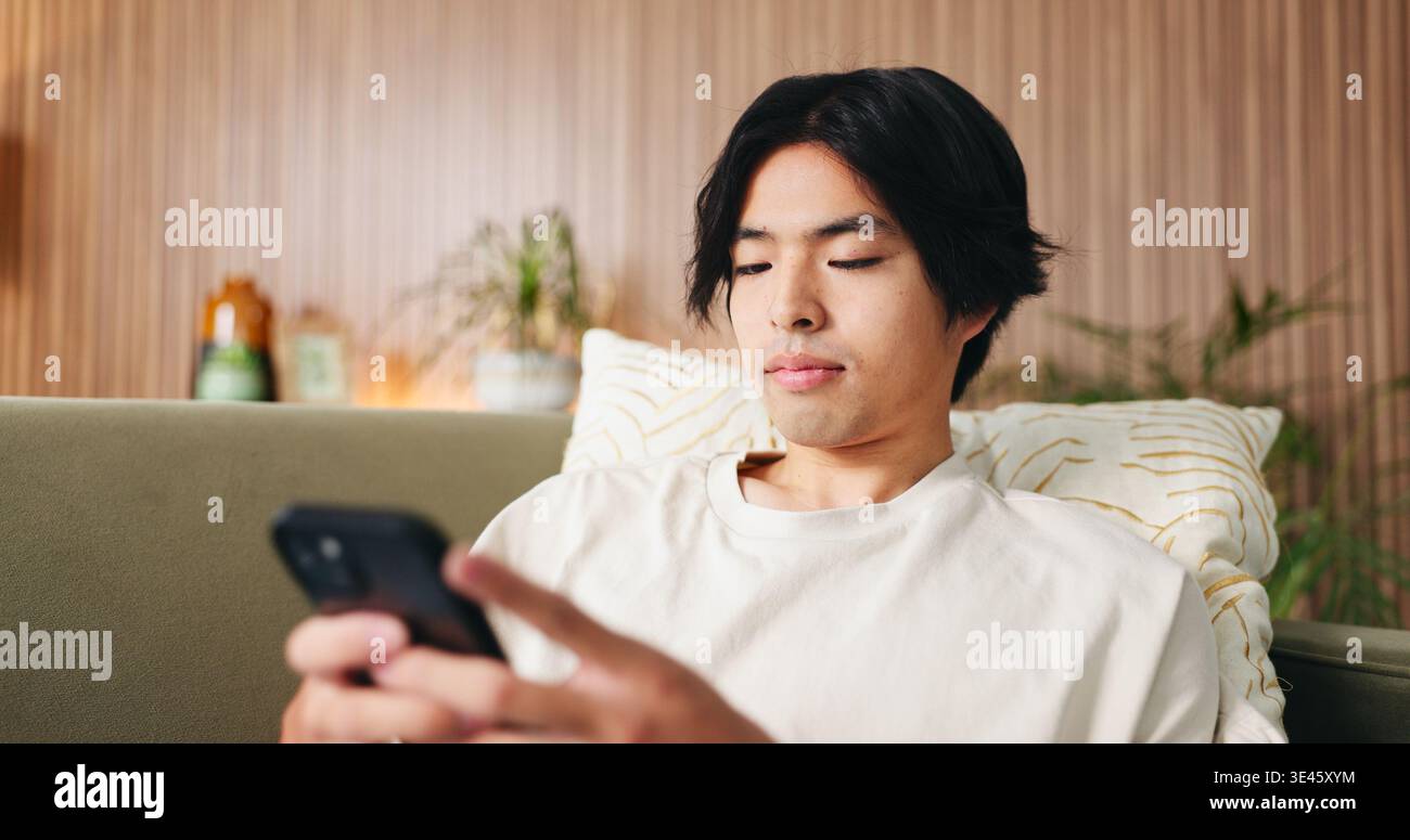 Lesen, Text und asiatischer Mann auf Couch mit Telefon, Internetsuche und Kommunikation auf Online-Plattform. Scrollen, digital und Person in House mit Technik Stockfoto