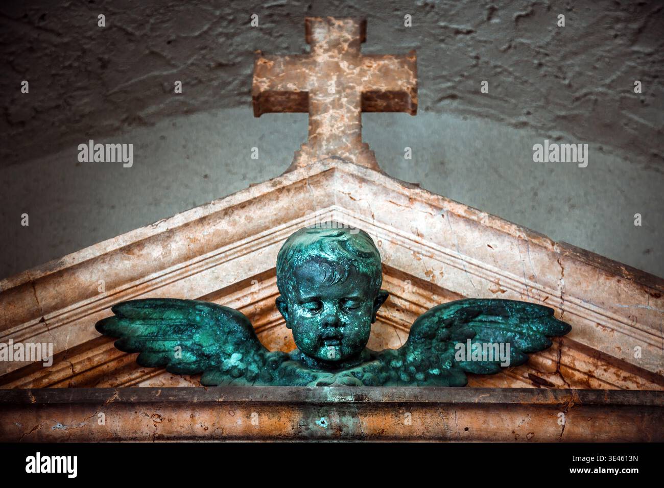 Eine verwitterte, detaillierte geflügelte Cherub-Statue im klassischen Stil befindet sich unter einem verzierten, moosbedeckten Steinkreuz. Die Skulptur ist gegen einen gerichtet Stockfoto