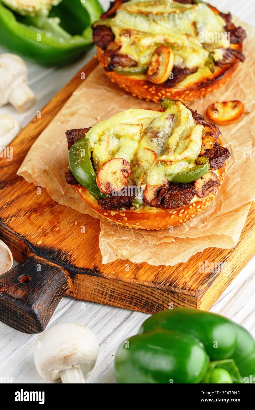 Sandwiches auf Sesambrötchen mit Rindfleisch, Paprika, Zwiebeln, Pilzen und Käse auf einem Holzbrett auf weißem Hintergrund. Rustikaler styl Stockfoto