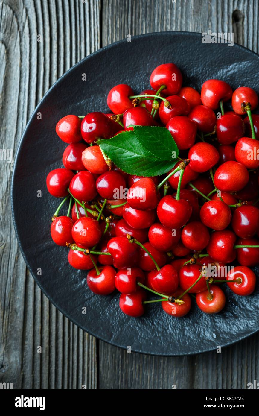 Blick von oben auf Kirschen, die auf einem schwarzen Teller auf einem Holztisch angeordnet sind. Frisches süßes Bio-Obst, minimales rustikales Design Stockfoto
