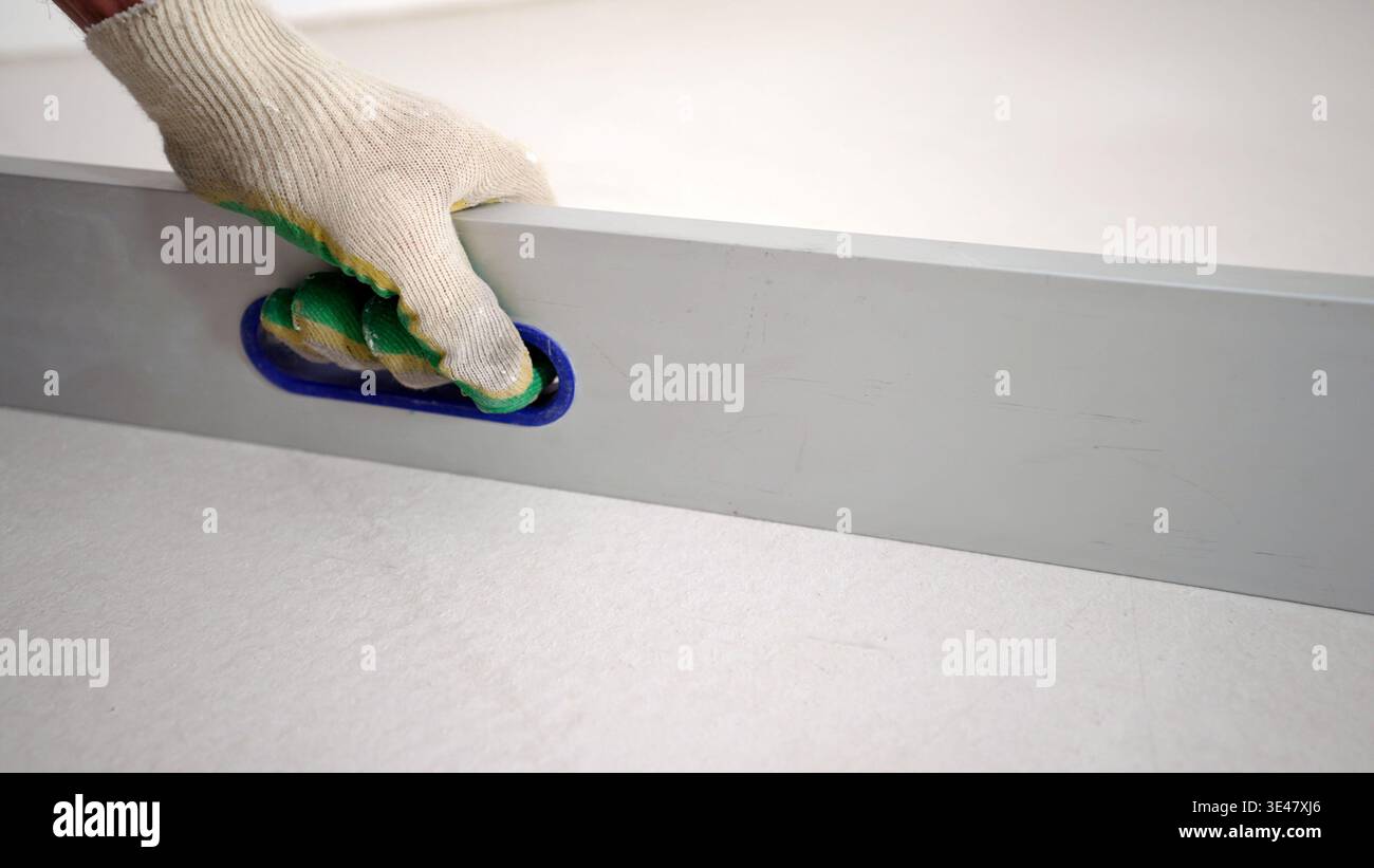 Der Baumeister in einem Arbeitshandschuh verwendet ein Wasserwaage-Werkzeug an einer frischen weißen Gipswand. Stockfoto
