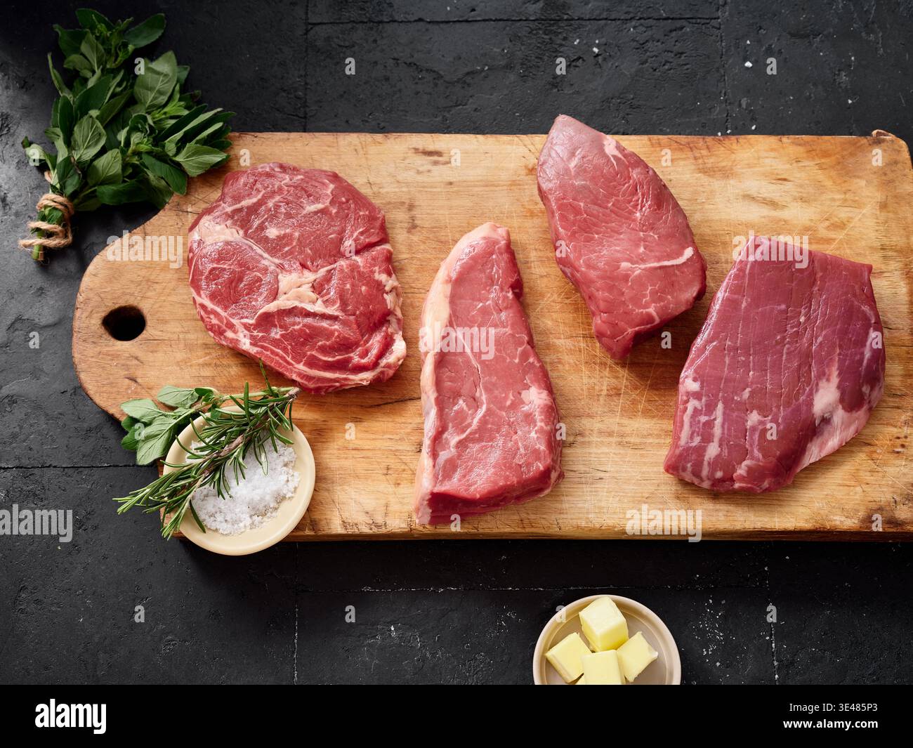 Mehrere Teile von rohem Fleisch werden auf ein hölzernes Schneidebrett gelegt. Zu den Aufschnitten gehören Rib-Eye, Roastbeef, Flanke und Rumpsteak. Frische Kräuter und Butter sind das Stockfoto