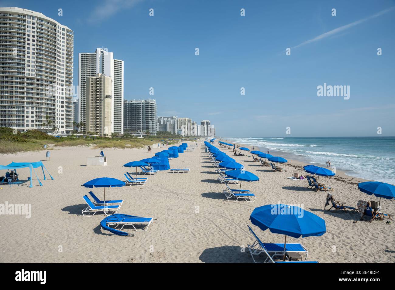 Riviera Beach, Florida, USA – 26. März 2026: Strand mit blauen Sonnenschirmen, Meereswellen und Hochhäusern entlang der Atlantikküste. Stockfoto