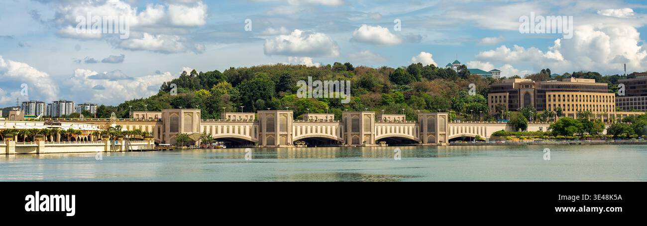 Panoramablick auf die Seri Gemilang-Brücke in Putrajaya, Malaysia, über ruhiges Wasser mit üppigen grünen Hügeln, moderner Architektur und einem bewölkten blauen Himmel Stockfoto